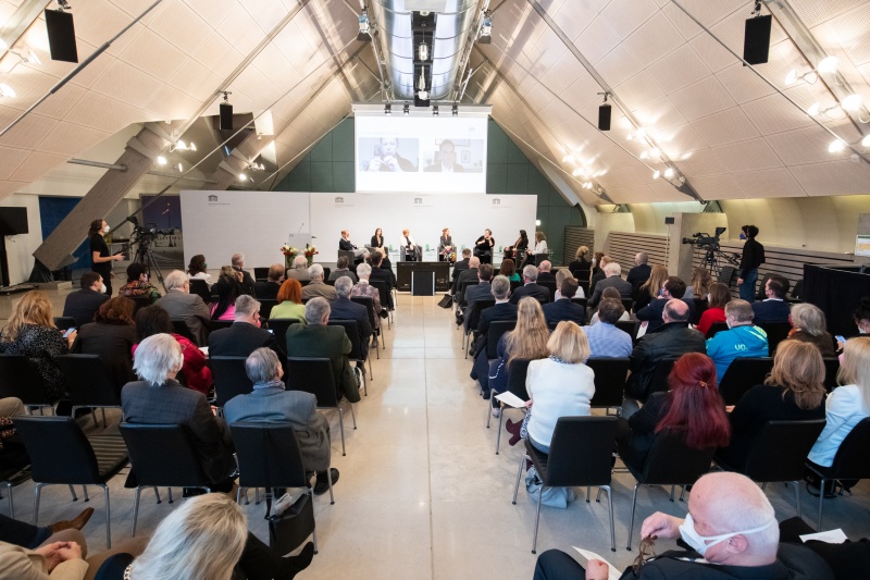 Blick in den Saal Richtung Bühne. Podiumsgespräch, von links: Moderator Universitätsprofessor Florian Bieber, Universität Graz, Nationalratsabgeordnete Ewa Ernst-Dziedzic (GRÜNE), Nationalratsabgeordnete Bedrana Ribo (GRÜNE), Nationalratsabgeordnete Carmen Jeitler-Cincelli (ÖVP), Nationalratsabgeordnete Eva Blimlinger (GRÜNE), Nationalratsabgeordnete Gudrun Kugler (ÖVP), Moderatorin Aleksandra Tomanic. Zugeschaltet auf der Leinwand von links: Bundesrat Christian Buchmann (ÖVP), Professor Damir Kapidžić, Universität Sarajewo