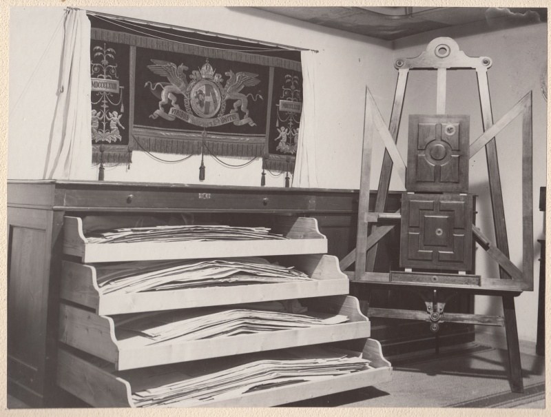 Das Bauplanarchiv des neuen Hauses, neueingerichtet vom technischen Assistenen Karl Albrecht. Oben: Der alte Brüstungsteppich der seinerzeitigen kaiserlichen Loge im Sitzungssaal, darunter ein Teil des Planarchivs, rechts auf einer Staffelei von Hansen zwei Brüstungsteile der Ministerbank des ehemaligen Abgeordnetenhauses mit den Einschüssen des Attentats auf den Justizminister Hohenburger am 5. Oktober 1911 (Foto veröffentlicht in der Zeitung 'Der Abend' am 2. Dez. 1933, S.8)