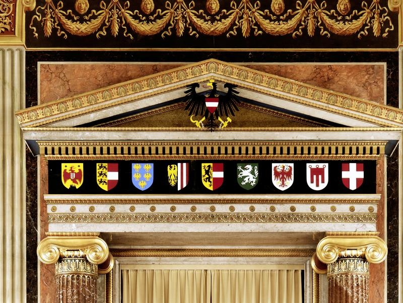 Ionische Säulen, darüber das Gebälk und Giebel mit dem Bundeswappen. Das Hauptfries des Gebälks zeigt die Landeswappen von Burgenland, Kärnten, Niederösterreich, Oberösterreich, Salzburg, Steiermark, Tirol, Vorarlberg und Wien (von li nach re). Ganz oben zu sehen ist ein Girlandenfries mit Tierköpfen.