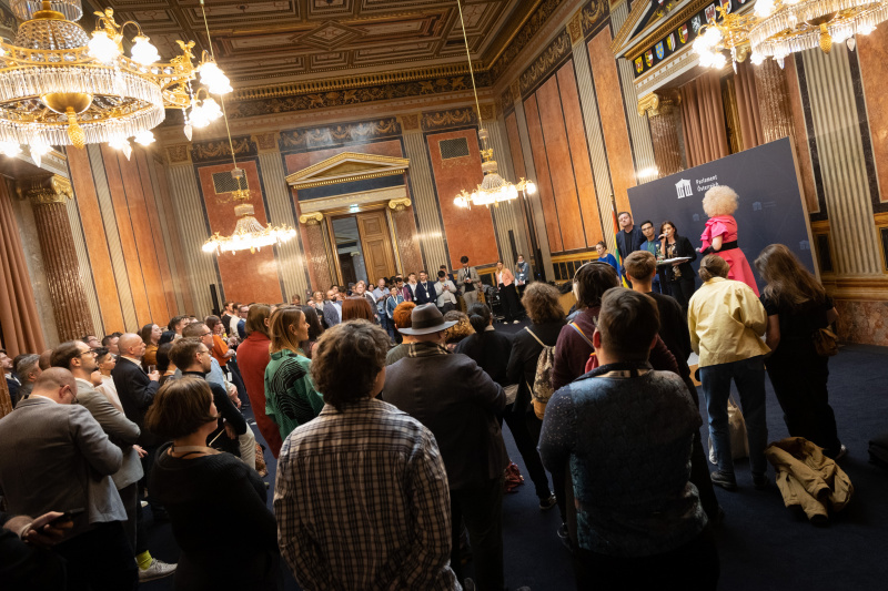 Blick in den Veranstaltungssaal, auf der Bühne die Steuerungsgruppe der LGBTIQ- Intergroup und Dragqueen Grazia Patricia