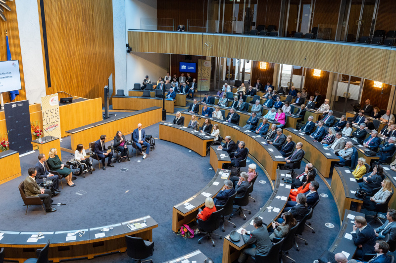 Blick Richtung Veranstaltungsteilnehmer:innen. Podiumsdiskussion, von links: Spitzensportler Pepo Puch, Spitzensportler Roman Rabl, Generalsekretärin des Österreichischen Paralympischen Committees Petra Huber, Moderatorin  Miriam Labus, Moderator Andreas Onea, Spitzensportlerin Claudia Lösch, Spitzensportler Florian Brungraber