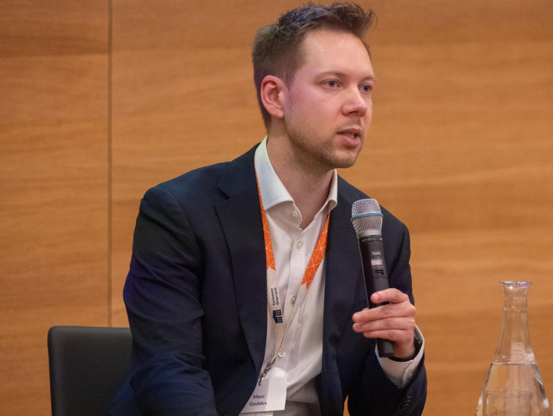 Marc Geddes sitzt am Podium und spricht ins Mikrofon.