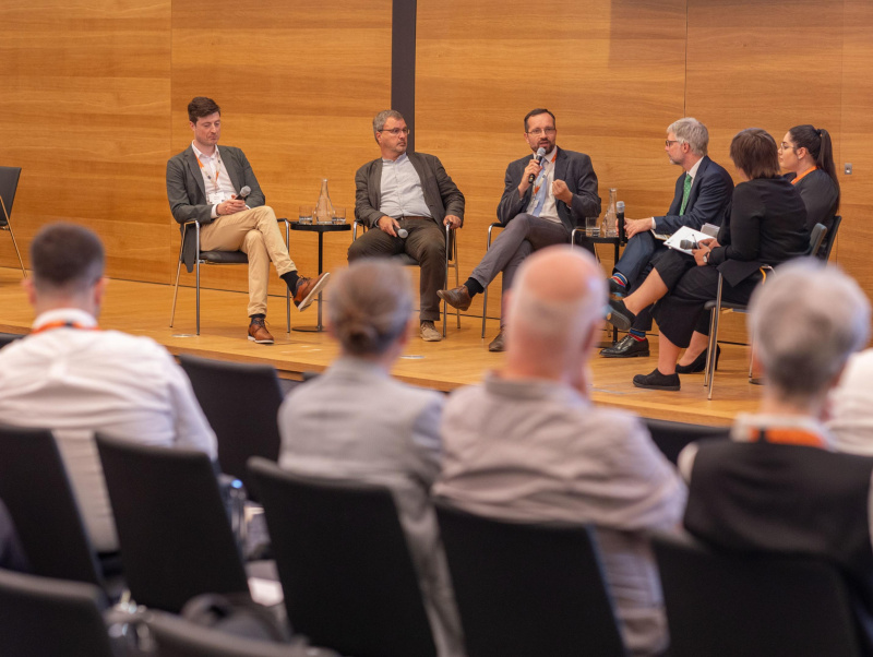 5 Teilnehmer:innen und Moderator des Panel 1 sitzen am Podium und blicken zu einer Teilnehmerin (Teresa Weber), die spricht.