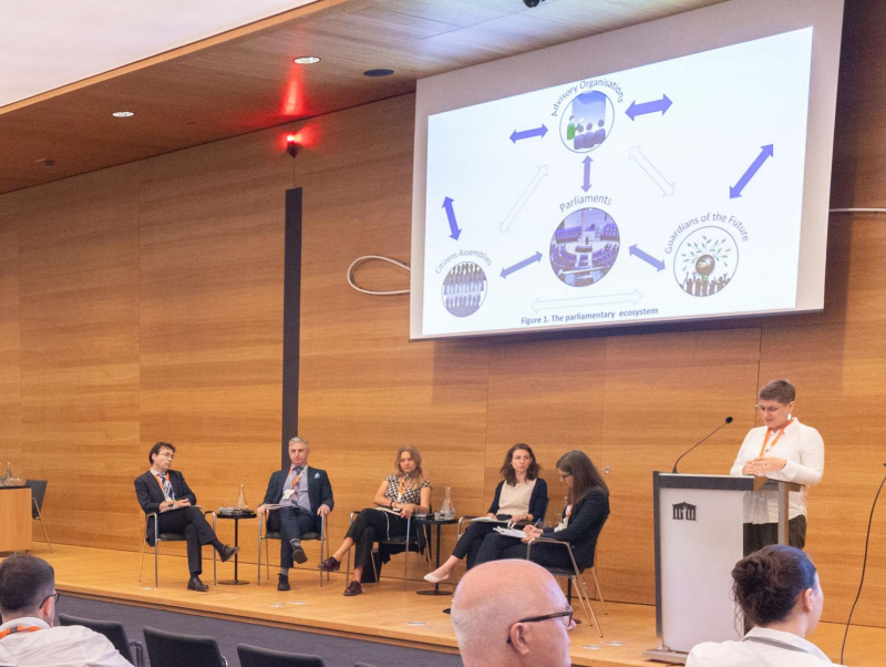 Teilnehmer:innen und die Moderatorin des Panel 3 sitzen am Podium und blicken zu einer Teilnehmerin (Sarah Nash), die am Rednerpult steht und vorträgt.