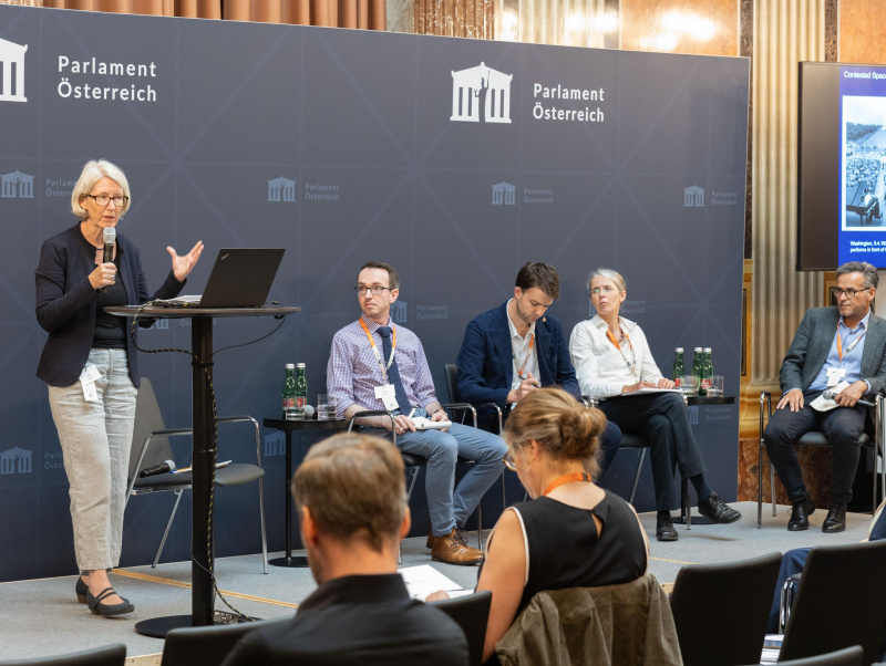 3 Teilnehmer:innen und der Moderator des Panel 4 sitzen am Podium und blicken zu einer Teilnehmerin (Anna Minta), die am Rednerpult steht und vorträgt.