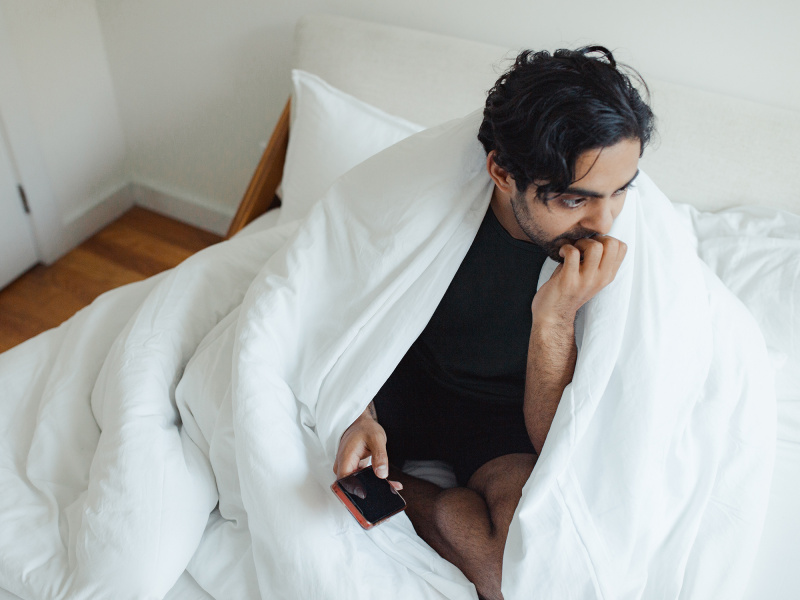 Offensichtlich verzweifelter Mann sitzt im Bett, Decke um die Schultern, in der Hand ein Smartphone, das nicht funktioniert
