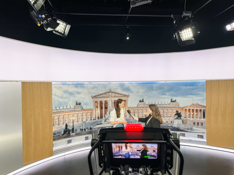 Im Foto ist der Bildschirm einer Kamera zu sehen, die zwei Frauen filmt. Die Rechtsstehende interviewt die Linksstehende. Das ganze findet in einem Studio statt. Im Hintergrund ist das Parlament zu sehen. 