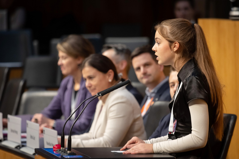 Schülerin am Redner:innenpult. Regierungsbank von links: Nationalratsabgeordneter Elisabeth Götze (GRÜNE), Nationalratsabgeordneter Christoph Pramhofer (NEOS), Nationalratsabgeordneter Bernhard Herzog (SPÖ), Nationalratsabgeordnete Bettina Zopf (ÖVP), Nationalratsabgeordneter Maximilian Weinzierl (FPÖ)