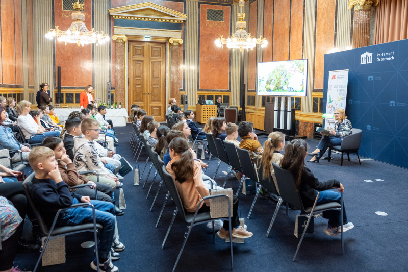 Blick Richtung Veranstaltungsteilnehmer:innen. Dritte Nationalratspräsidentin Doris Bures (SPÖ) liest Kindern aus einem Buch vor