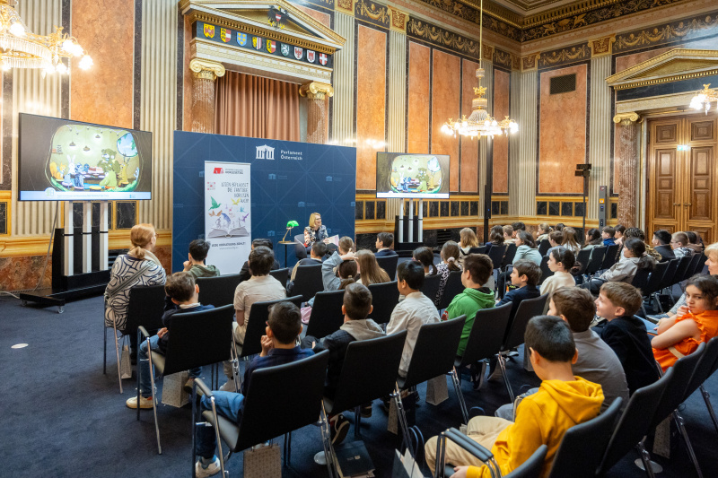 Blick Richtung Veranstaltungsteilnehmer:innen. Dritte Nationalratspräsidentin Doris Bures (SPÖ) liest Kindern aus einem Buch vor.
