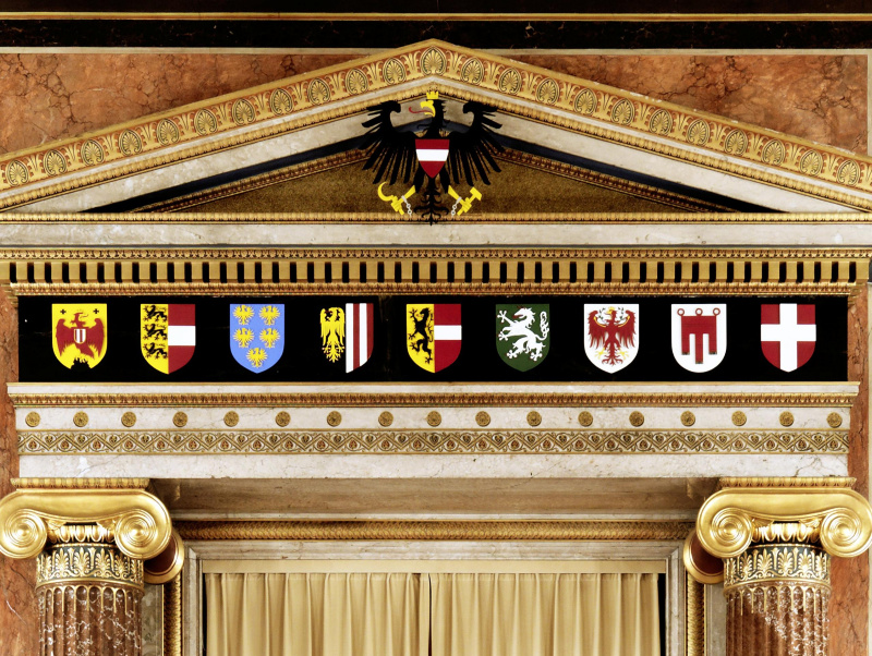 Ionische Säulen, darüber das Gebälk und Giebel mit dem Bundeswappen. Das Hauptfries des Gebälks zeigt die Landeswappen von Burgenland, Kärnten, Niederösterreich, Oberösterreich, Salzburg, Steiermark, Tirol, Vorarlberg und Wien (von li nach re). Ganz oben zu sehen ist ein Girlandenfries mit Tierköpfen.