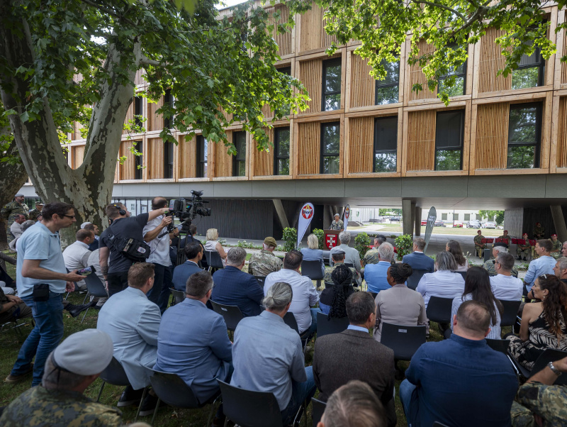 Menschen sitzen beim Festakt am Boden - im Hintergrund ein Redner am Pult und das neue Unterkunftsgebäude in moderner Holzbauweise