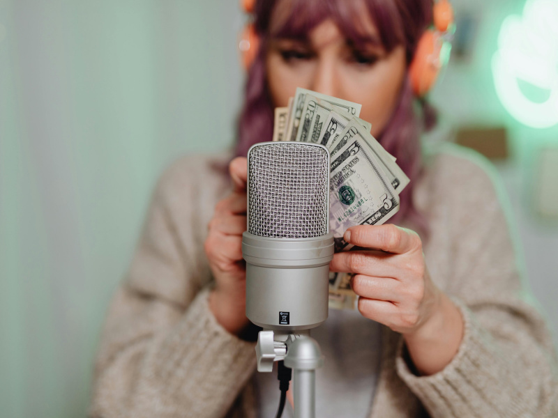 Eine Frau mit aufgesetzten Kopfhörern hält Geldscheine vor ein Podcast-Mikrofon.