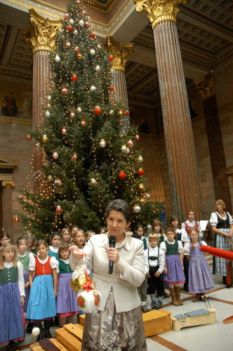 Barbara Prammer am Mikrofon, sie hält eine Christbaumkugel, im Hintergrund der Kinderchor vor dem Weihnachtsbaum.