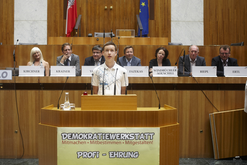 Schüler der zum Demokratiewerkstatt-Profi geehrt wird hält eine kurze Rede. Im Hintergrund auf der Regierungsbank v. links: Elke Kirchner - Klub der Freiheitlichen, Kai Jan Krainer - Nationalratsabgeordneter, Bernhard Vock - Nationalratsabgeordneter, Hannes Fazekas - Nationalratsabgeordneter, Tanja Windbüchler-Souschill - Nationalratsabgeordnete, Franz Riepl - Nationalratsabgeordneter und Bundesratsmitglied Johann Ertl