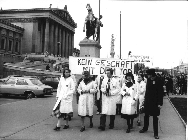 DemonstrantInnen in weißen Arztmänteln vor dem Parlament protestieren gegen die Straffreiheit des Schwangerschaftsabbruchs ("Fristenlösung"). Sie tragen ein Transparent mit der Aufschrift "Kein Geschäft mit dem Tod". Links im Hintergrund das Parlament mit Rampe.