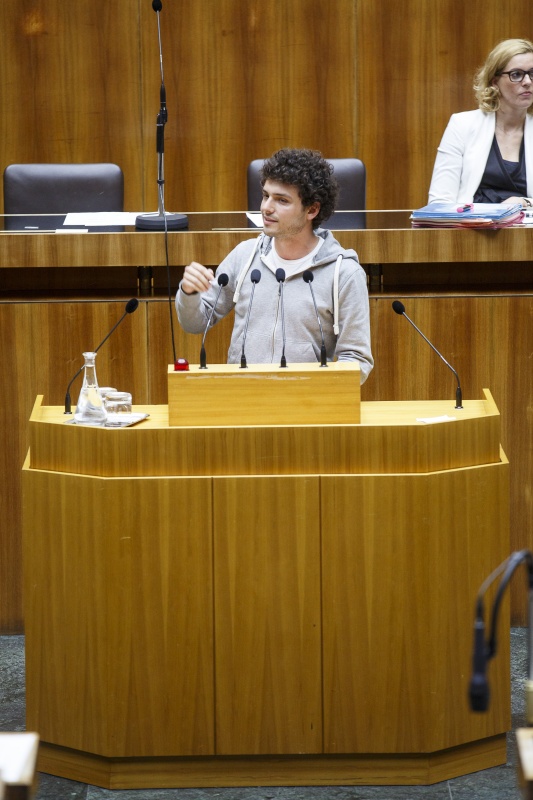Nationalratsabgeordneter Julian Schmid (G) am Rednerpult. Im Hintergrund auf der Regierungsbank Staatssekretärin Sonja Steßl
