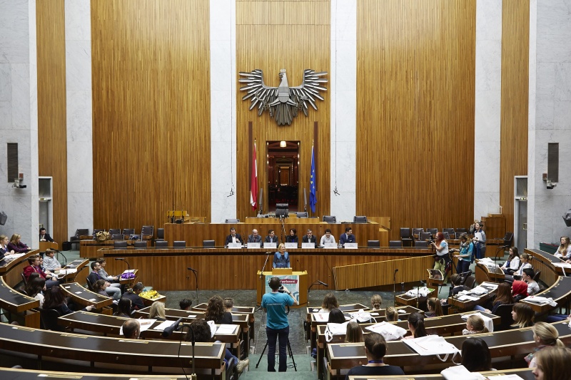 Nationalratspräsidentin Doris Bures (S) begrüßt die SchülerInnen. Am Podium v.li.: Die Nationalratsabgeordneten Robert Lugar (T), Christoph Vavrik (N) und Asdin El Habbassi (V), Bundesratspräsidentin Ana Blatnik (S) sowie die Nationalratsabgeordneten Hermann Lipitsch (S), Julian Schmid (G) und Philipp Schrangl (F). Blick Richtung VeranstaltungsteilnehmerInnen